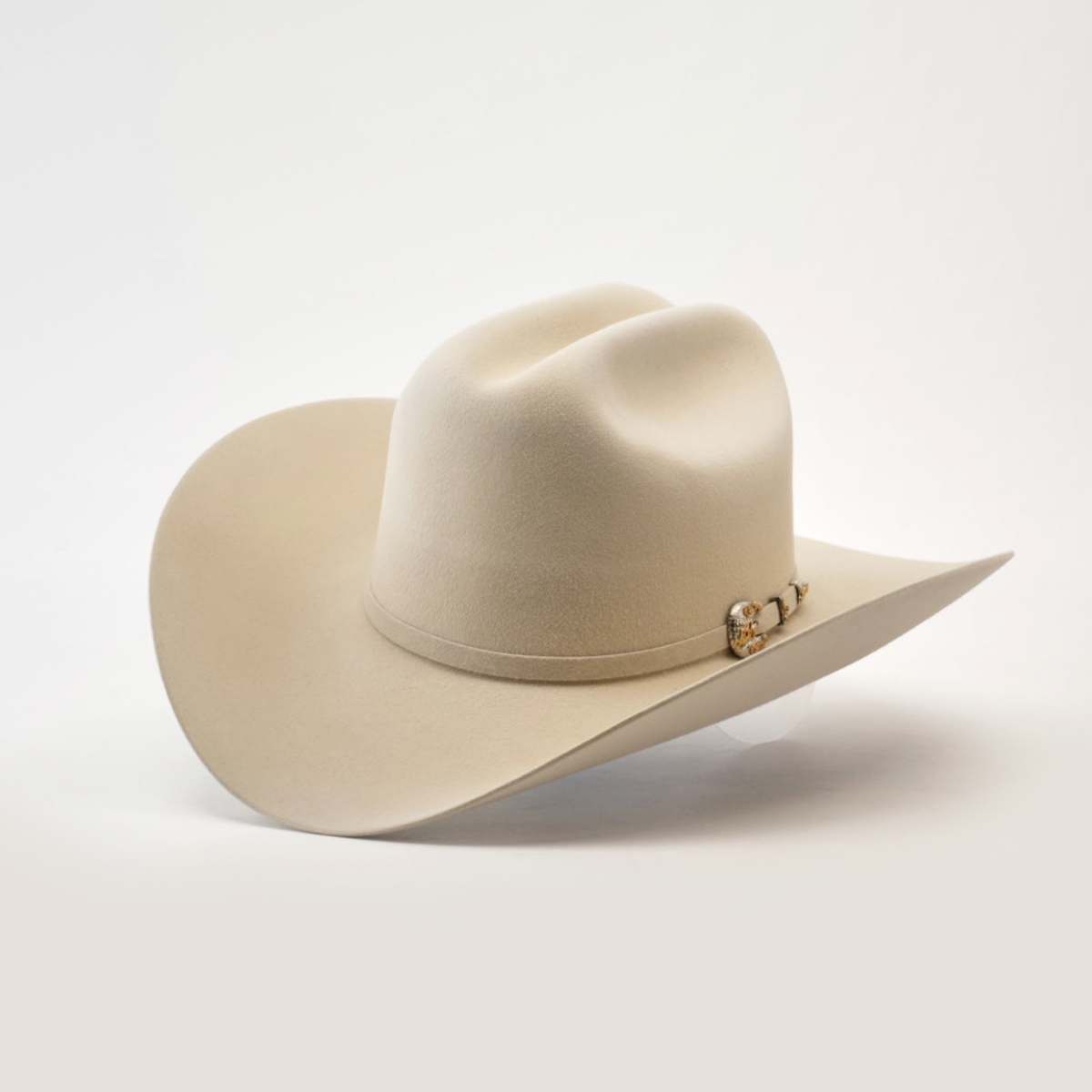 The 1000X Imperial Belly hat is a beige cowboy style with a wide brim, adorned with a decorative band featuring a small metallic emblem, set against a plain white background.