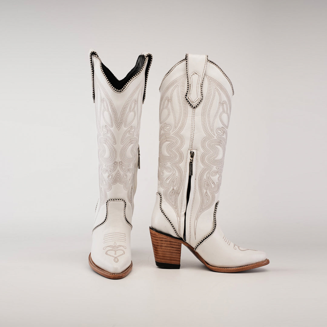 The Linda Studs Tall Shaft Frost White - J Toe cowboy boots, part of the Platinum Collection, boast artisanal craftsmanship with intricate embroidery. Made from premium leather, these stylish boots feature pointed toes, a side zipper, and stacked wooden heels against a plain background.