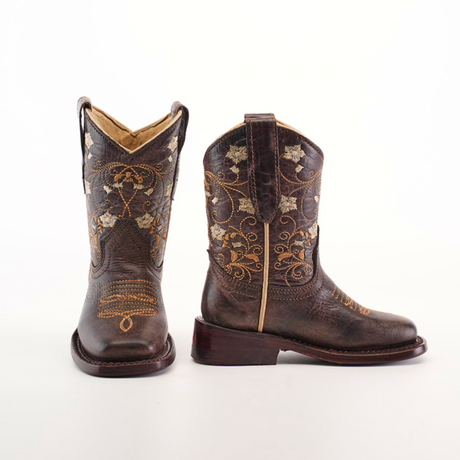 The Dariela Chocolate boots for girls boast premium leather with a square toe and intricate gold floral embroidery. One boot stands upright to showcase the detailed design, while the other is angled slightly on a plain white background.