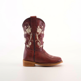 The Jatziry Cogñac Square Toe boot for little girls features intricate white floral embroidery on handcrafted leather, a pull strap, and a wooden heel—an ideal choice for budding cowgirls.