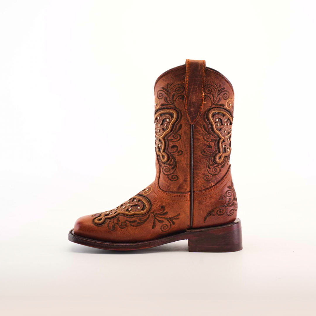 The Butterfly Studs Cogñac boot features a single brown cowhide leather design with intricate embroidery, butterfly studs, swirls, and floral patterns. It has a rounded toe, pull strap, low wooden heel, all displayed against a plain white background.