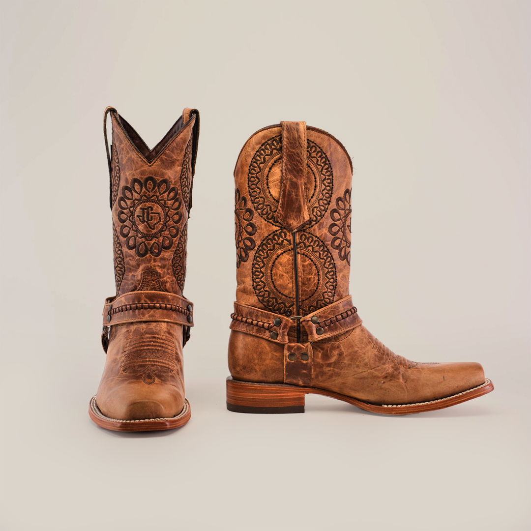 The luxurious Siberia Fawn - Biker Toe cowboy boots are crafted from premium cowhide, featuring intricate floral patterns and decorative stitching. A sturdy heel and a strap with metal accents across the vamp enhance their elegance, set against a plain white background.