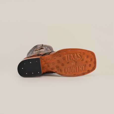 Bottom view of the Pirarucu Print Tan - Square Toe cowboy boot with TEXAS COUNTRY embossed on the premium cowhide sole. The heel has metal studs and decorative patterns for an artisan flair, all set against a plain white background.