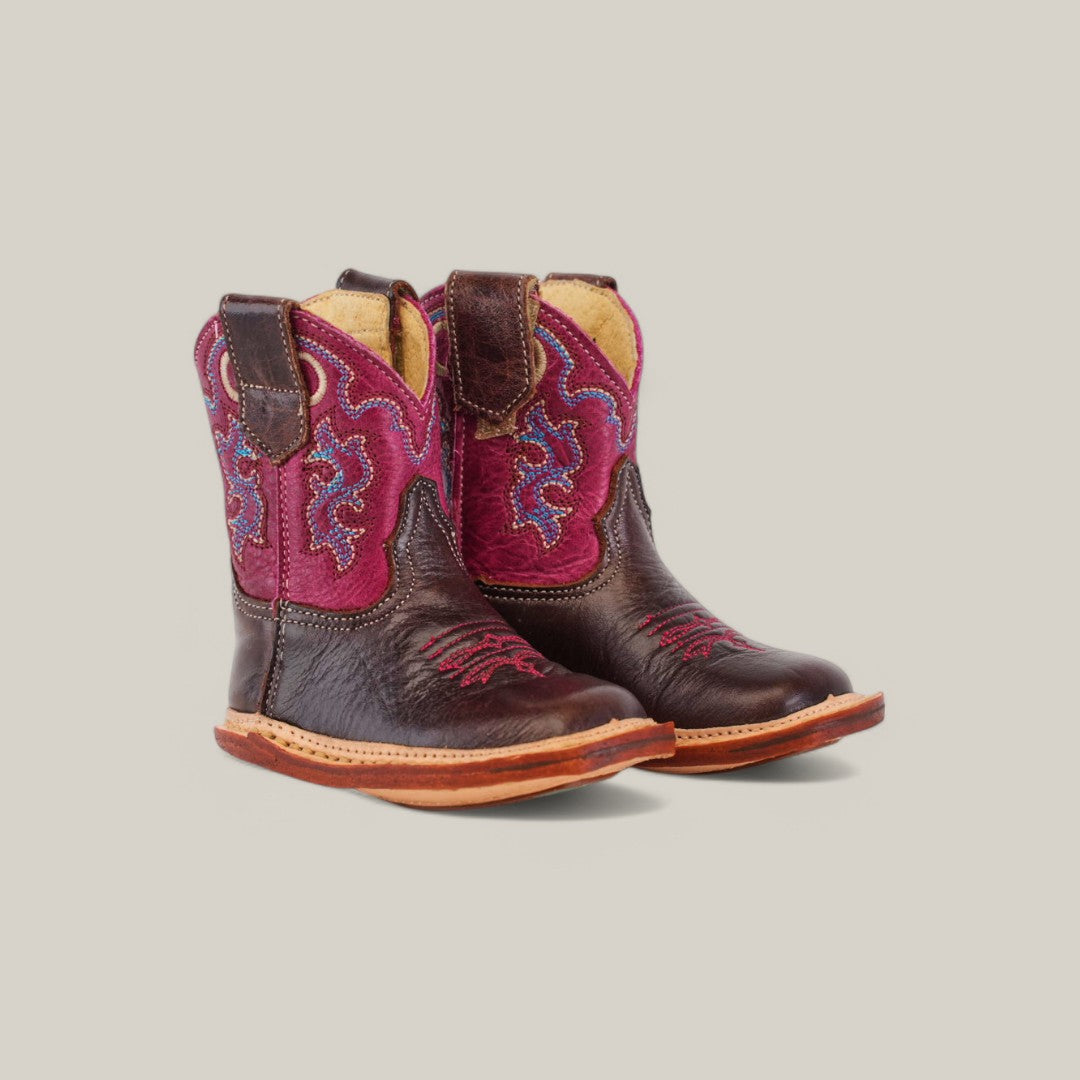 The Emma Baby Boots, featuring a brown leather and vibrant pink upper with blue embroidery, include a comfortable square toe and sturdy wooden soles. Set against a light gray background, theyre the perfect mix of style and practicality for your little adventurer.