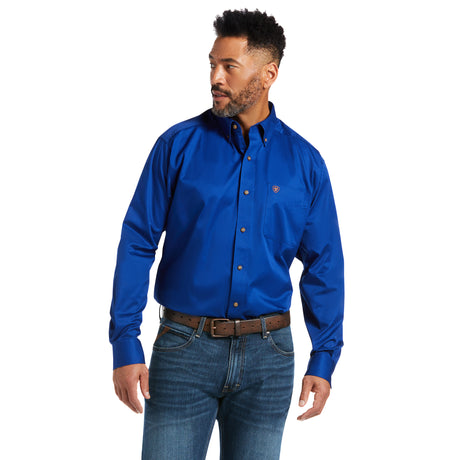 A man with short curly hair and a beard wearing a Solid Twill Classic Fit Shirt Blue - 10006660 and blue jeans with a brown belt stands looking to his right. The 100% Cotton Twill shirt showcases a small logo on the chest.