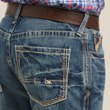 A person wearing M4 Low Rise Boundary Boot Cut Jeans in Gulch with visible stitching and a brown leather belt. The top is a partially visible red and blue plaid shirt, and the jeans feature back pockets with contrast stitching.