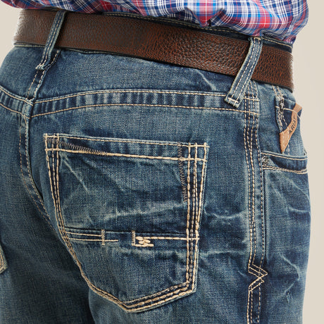 A person wearing M4 Low Rise Boundary Boot Cut Jeans in Gulch with visible stitching and a brown leather belt. The top is a partially visible red and blue plaid shirt, and the jeans feature back pockets with contrast stitching.