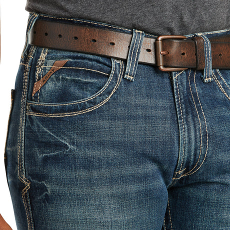 Close-up of a person wearing M5 Slim Boundary Stackable Straight Leg Jeans, Gulch - 10014010, featuring blue denim with contrast stitching, a small pocket with leather tag, paired with a brown leather belt and dark gray shirt.