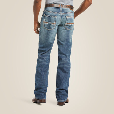 A person in M4 Low Rise Coltrane Boot Cut Jeans - Durango stands with their back to the camera. The denim has two stitched back pockets and is paired with a brown belt and shoes, set against an off-white background.