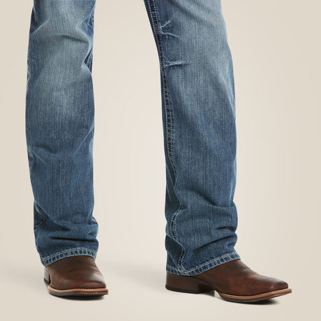 A close-up of a persons lower legs showcases the M4 Low Rise Coltrane Boot Cut Jean - Durango, paired with brown leather cowboy boots. The jeans feature heavy-duty cotton denim with a modern boot cut, slight fading, and distressing near the knees against a light background.