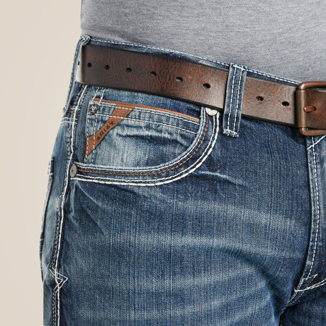 A person is wearing M4 Low Rise Coltrane Boot Cut Jeans - Durango, featuring heavy-duty blue denim with white stitching and a small brown label near the front pocket. A brown leather belt and tucked-in gray shirt complete the modern look.