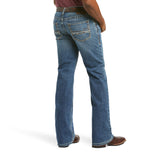 A person is wearing M5 Slim Stretch Stillwell Stackable Straight Leg Jean - Fargo, featuring premium cotton in blue with brown stitching, paired with a dark belt and brown shoes. The image shows the back view from the waist down against a plain white background, highlighting two decorative-stitched back pockets.