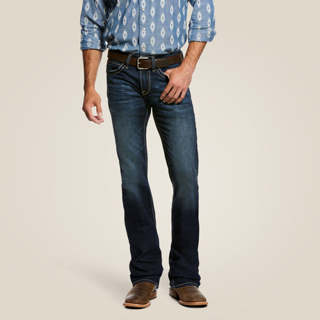 A person wears a blue patterned shirt, dark blue M7 Rocker Concord Stretch Stackable Straight Leg Jean - Dodge - 10026681 by Ariat, a black belt, and brown boots against a neutral background. Their hand rests on their hip, highlighting jeans with a faded thigh effect and stretch fabric.