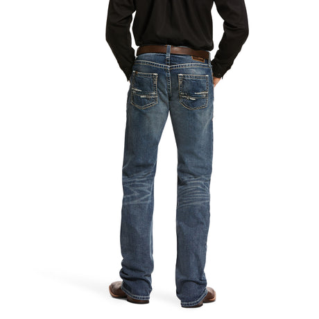 A person wears the M5 Slim Stretch Adkins Stackable Straight Leg Jean (lennox - 10030275), featuring durable denim with a faded look and decorative stitching on the back pockets, complemented by a brown belt and shoes. Theyre in a black shirt against a plain white background.