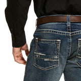 A person is wearing M5 Slim Stretch Adkins Stackable Straight Leg Jean (lennox - 10030275) with frayed back pockets, a brown leather belt, and a black shirt with buttoned cuffs. Their left hand rests by their side, highlighting the jeans durable denim and stylish worn look.