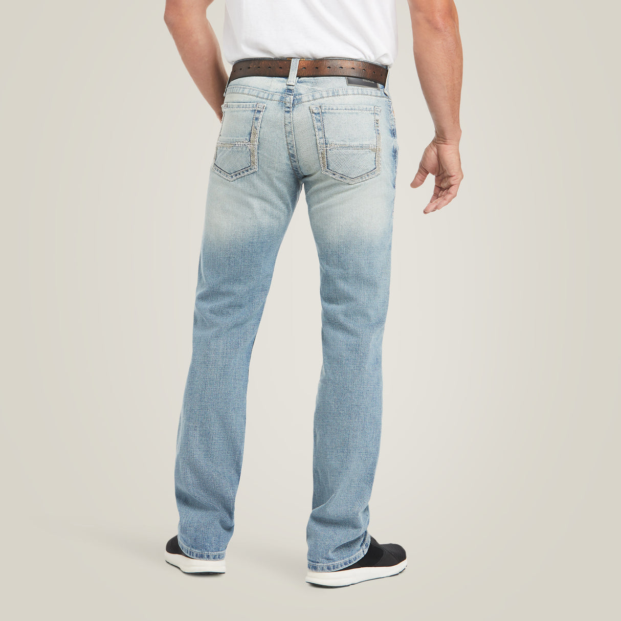 Rear view of a person in M7 Rocker Stretch Stirling Stackable Straight Leg Jeans - Shasta, displaying light blue faded stretch fabric. They wear a brown belt and white shirt, stand with one leg bent in black and white sneakers, emphasizing the relaxed straight-leg fit.