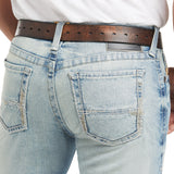 The person is wearing the M7 Rocker Stretch Stirling Stackable Straight Leg Jean - Shasta, light blue with distressed, frayed back pockets. Paired with a brown leather belt and a crisp white shirt tucked in, these jeans offer an effortlessly casual look.