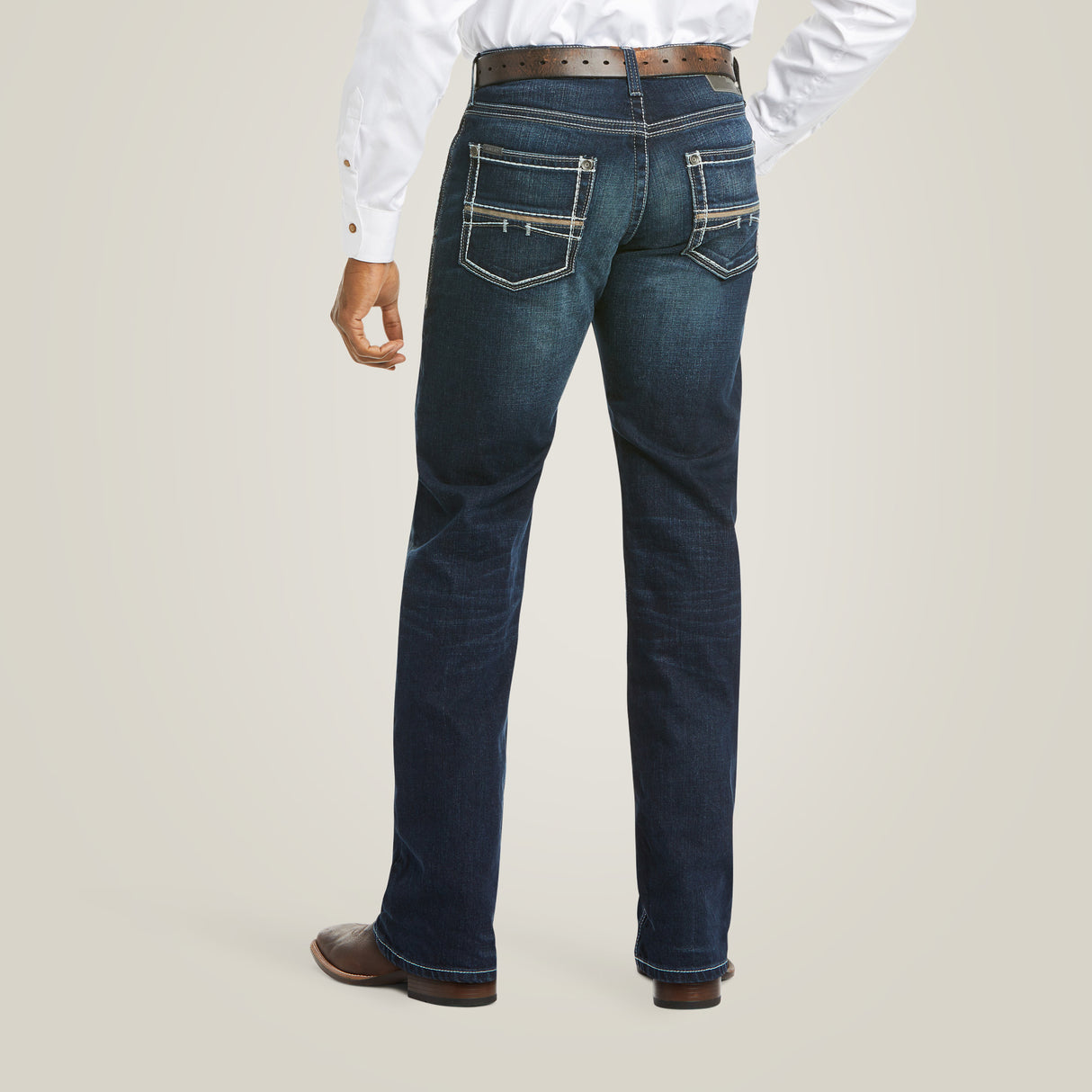 A person in a white shirt and brown belt is wearing the M5 Slim Stretch Coltrane Stackable Straight Leg Jean in Nightingale, featuring decorative stitching and reinforced pockets, standing against a light, neutral background.