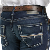 Close-up of a person wearing M5 Slim Stretch Coltrane Stackable Straight Leg Jean - Nightingale with white stitching and a brown leather belt. Reinforced pockets feature a DENIM metal tag above the back pocket, complementing the white shirt tucked in.