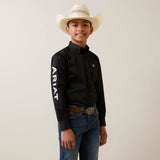 A young person in a cowboy hat wears the Ariat Boys Black & White Team Logo Embroidered Long Sleeve Western Shirt - 10045426, paired effortlessly with blue jeans. The wrinkle-resistant material keeps the black button-up crisp as they smile against a plain background.