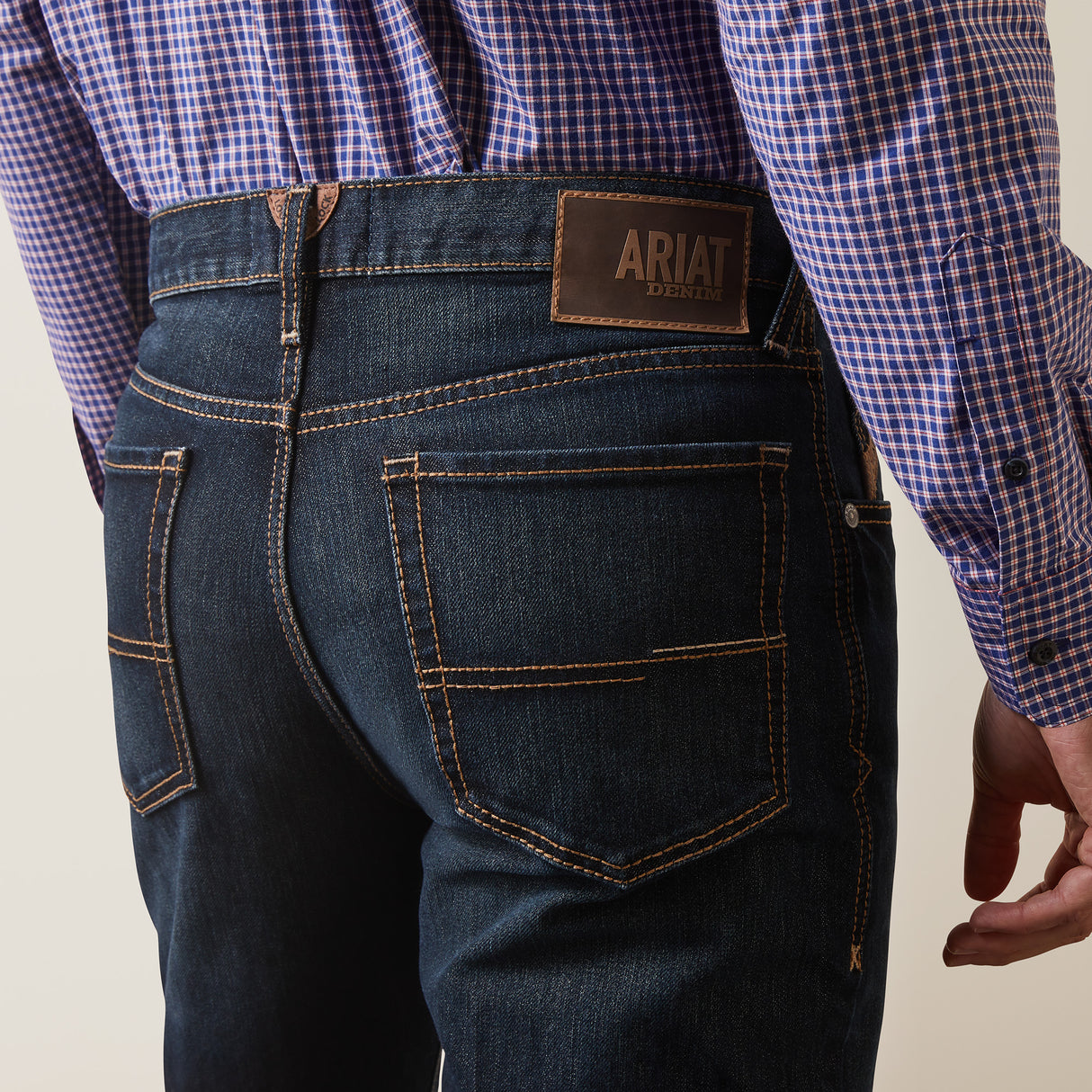 A person wearing a blue and purple checkered shirt and ARIAT M4 Relaxed Hansen Boot Cut Jeans in Blackstone, featuring dark blue denim with brown stitching and a visible leather patch labeled ARIAT DENIM on the back.