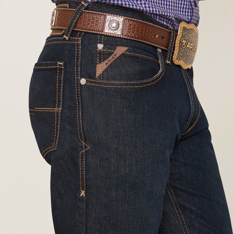 A person wears the M4 Relaxed Hansen Boot Cut Jean in Blackstone, featuring beige stitching and a brown leather belt with a decorative buckle. The jeans have a logo patch and stylized pockets, set against a plain white background.