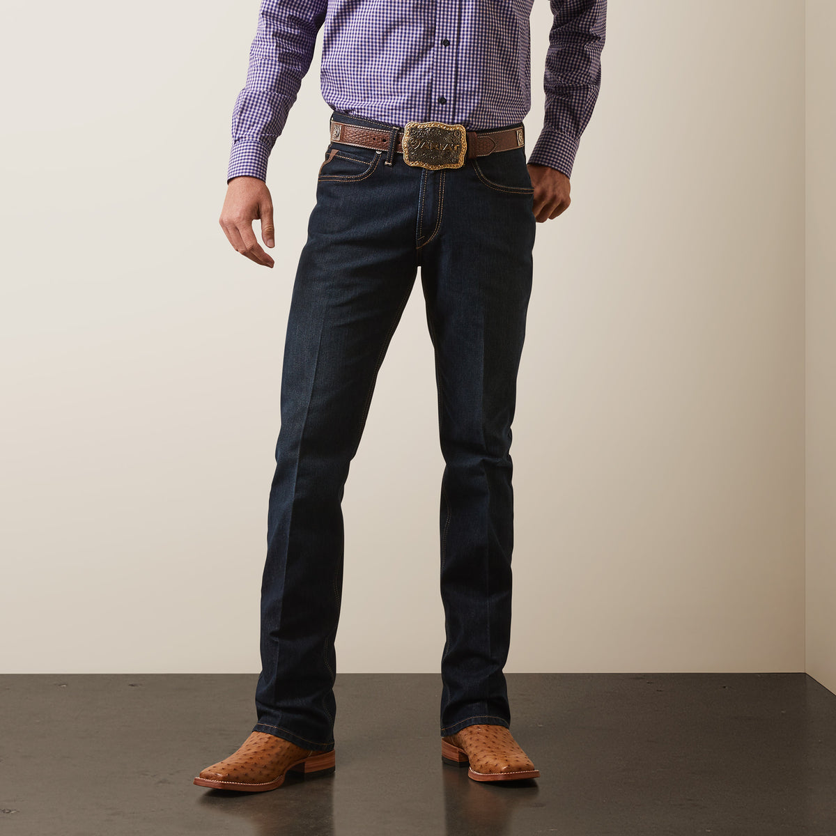 A man in a purple checkered shirt and dark blue M4 Relaxed Hansen Boot Cut Jean - Blackstone stands confidently on a textured floor. Hes wearing brown leather cowboy boots with a large belt buckle, focusing on the relaxed fit of his denim and the lower half of his body.