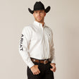 A man stands confidently against a plain background wearing the Team Logo Twill Classic Fit Shirt White (10046825), featuring ARIAT on the sleeve. He completes his look with a black cowboy hat, brown belt, and black pants, gazing to the side with his hand resting casually on his belt.