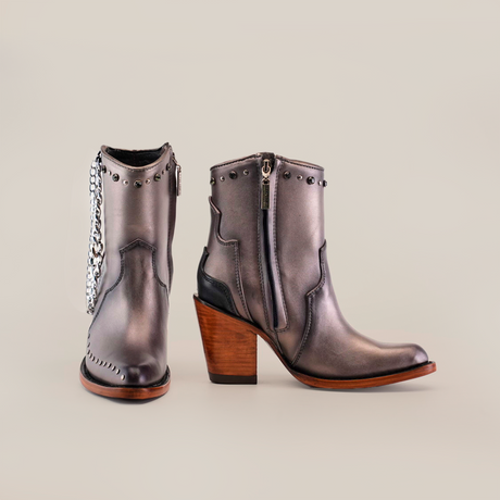 Introducing the Milan Chains Bootie Silver - J Toe: stylish gray ankle boots with decorative studs and a silver chain detail. Part of the Platinum Collection, these handcrafted boots feature side zippers and a chunky wooden heel, all set against a plain white background.