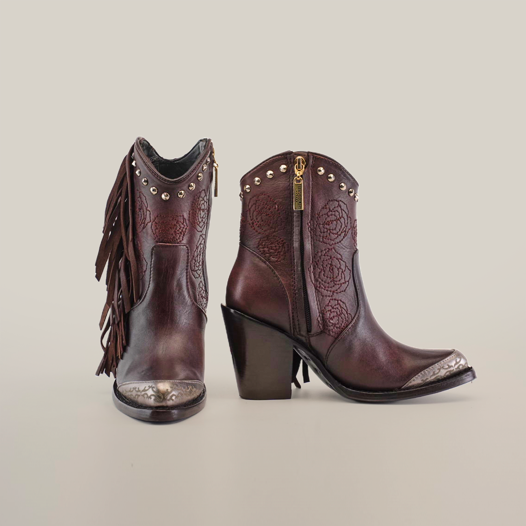 The Dallas Fringe Bootie Chocolate J Toe, from the Platinum Collection, features chocolate brown leather with floral embossing and silver-detailed toe caps. It includes side fringe on one boot and metallic studs near the top, set against a plain white background.