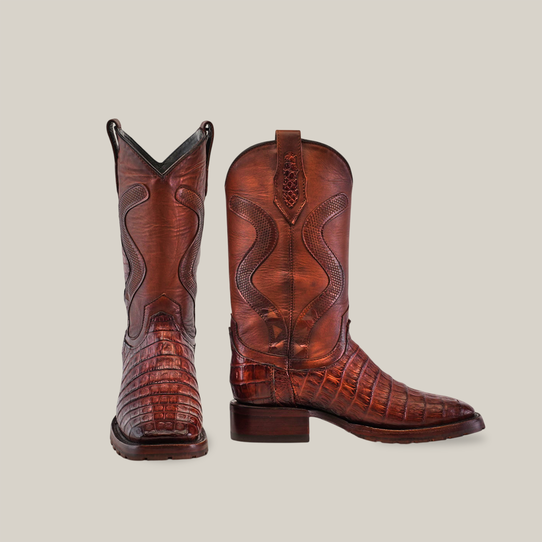 The Exotic Caiman Belly Tail - Diamond boots in brandy color capture Western fashions essence with an alligator skin pattern. One boot stands to show intricate stitching, while the other lies on its side, highlighting a square toe and double sole.