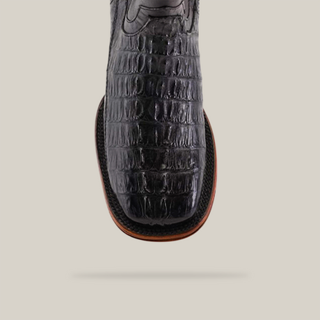 Close-up of the Exotic Caiman Tail - Diamond - Black cowboy boot with a square toe, featuring a black crocodile pattern. Its polished finish and brown sole add elegance against a plain light beige background.