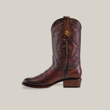 The Western - Platinum - Brown boot features decorative stitching, a logo on the pull strap, a slightly pointed roper toe, and a low wooden heel. Its showcased against a plain white background.