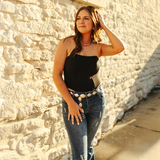 A woman with long hair poses against a sunlit stone wall, wearing the LB Black Jumper Corset and blue jeans with a decorative belt. She looks to the side, one hand on her hip and the other brushing her hair.