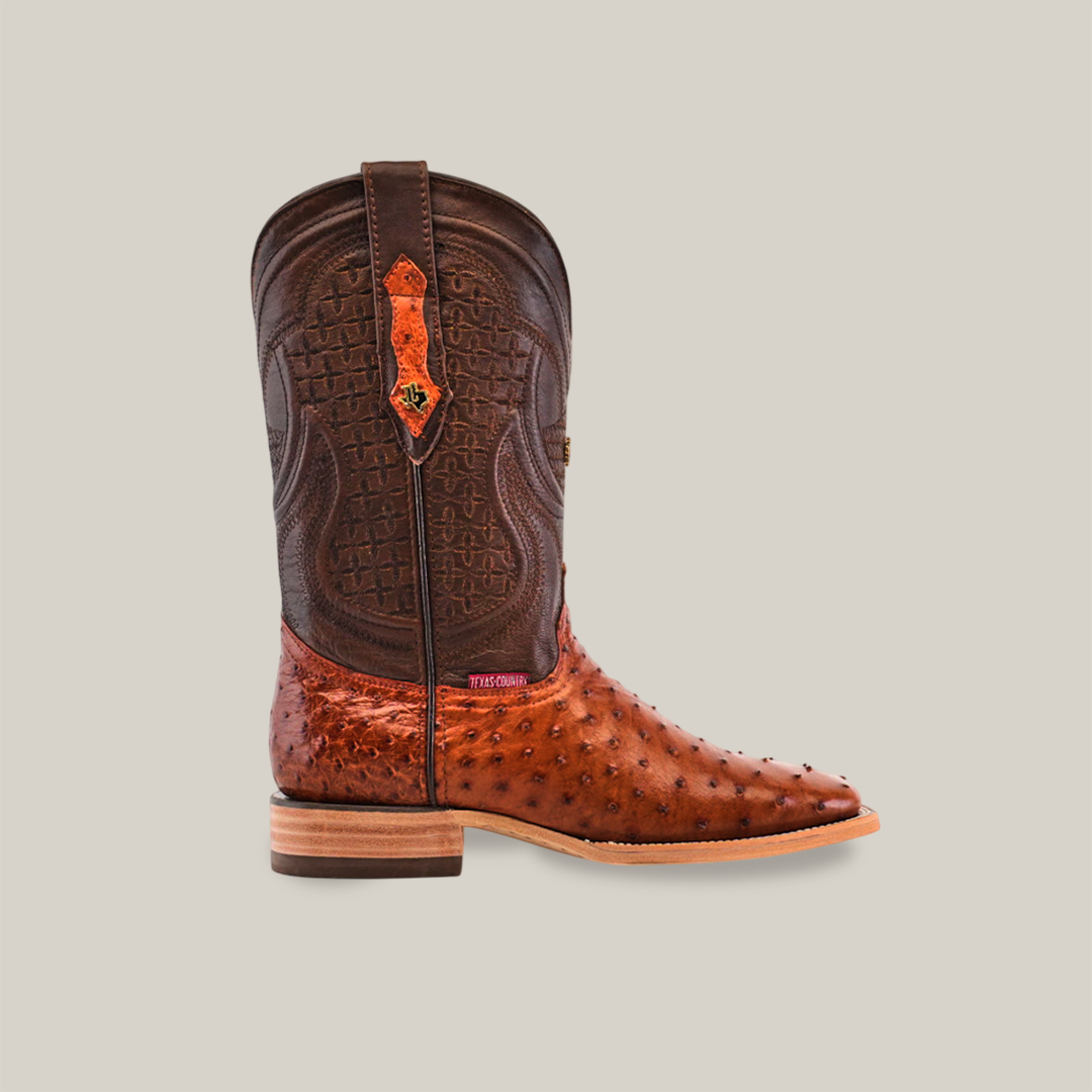 The Exotic Full Quilt Ostrich - Brandy - Square Toe boot features an intricate leather pattern on the shaft and unique ostrich texture on the foot, with a short wooden heel and decorative pull strap, embodying the cowboy lifestyle against a plain, light-colored backdrop.