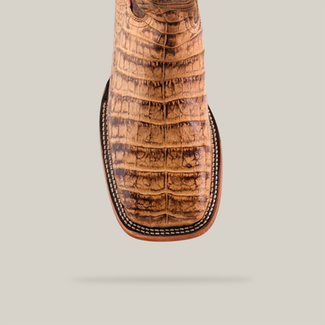 A close-up of the Exotic Caiman Belly - Antique - Square Toe boot highlights its brown square-toed crocodile leather with distinctive texture and intricate stitching. Ideal for cowboy fashion lovers, its set against a light background, casting a shadow beneath.