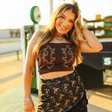 A smiling woman with long hair stands by a vintage gas pump, wearing the LB Brown Corset Cream Embroidery T0655-BRN, paired with a dark fringe skirt. The sun casts a warm glow on the charming scene.