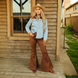 A woman in a LB Denim Camel Suede Pearl Snap TO718 shirt, brown flared pants, a wide-brimmed hat, and brown boots stands confidently on a wooden porch before a rustic building. A grassy area and another structure enhance the scenes charm.