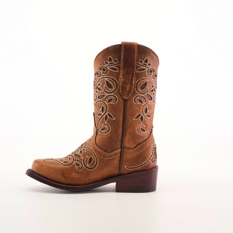 The Carola Tabaco - Snip Toe boot is a single brown cowboy boot made of premium leather, adorned with detailed white embroidery on the shaft and foot. It features a low heel and pull straps on either side, set against a simple white background.