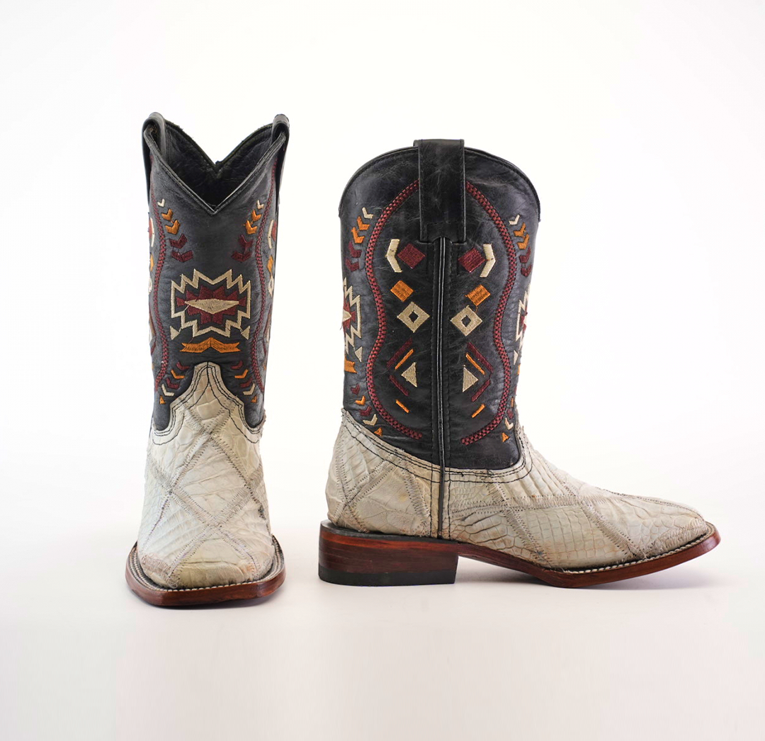 The Exotic American Alligator Patchwork Bone Square Toe cowboy boots flaunt a square toe and intricate red, orange, and beige tribal patterns on a black shaft. Its foot area displays textured design with light colors and patchwork details, completed with a wooden heel and sole.