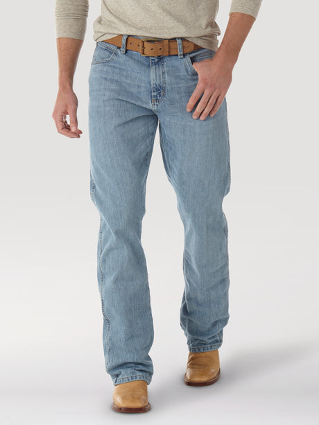 A person wearing Mens Wrangler Retro Relaxed Fit Bootcut Jeans - WRT20CR in light blue, a beige long-sleeve shirt, a brown leather belt, and tan boots stands against a plain white background.