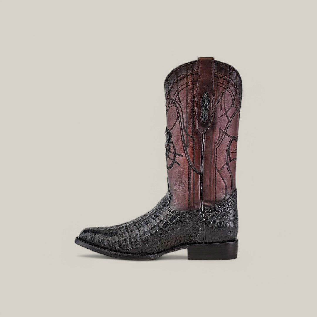 The Plunge Black Caiman Laser & Embroidery - Round Toe - CU422 cowboy boot features a dark brown upper with intricate stitching and a textured black Caiman leather lower. It has a pointed toe and low heel, all set on a plain light background.