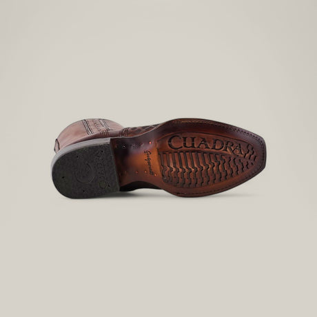 The image displays the sole of the Over Maple Laser & Woven - Narrow Square Toe - CU674 brown leather shoe, featuring intricate stitching and a woven texture. CUADRA is embossed on the sole, which has a textured grip pattern, while a black rubber section on the heel offers extra traction.