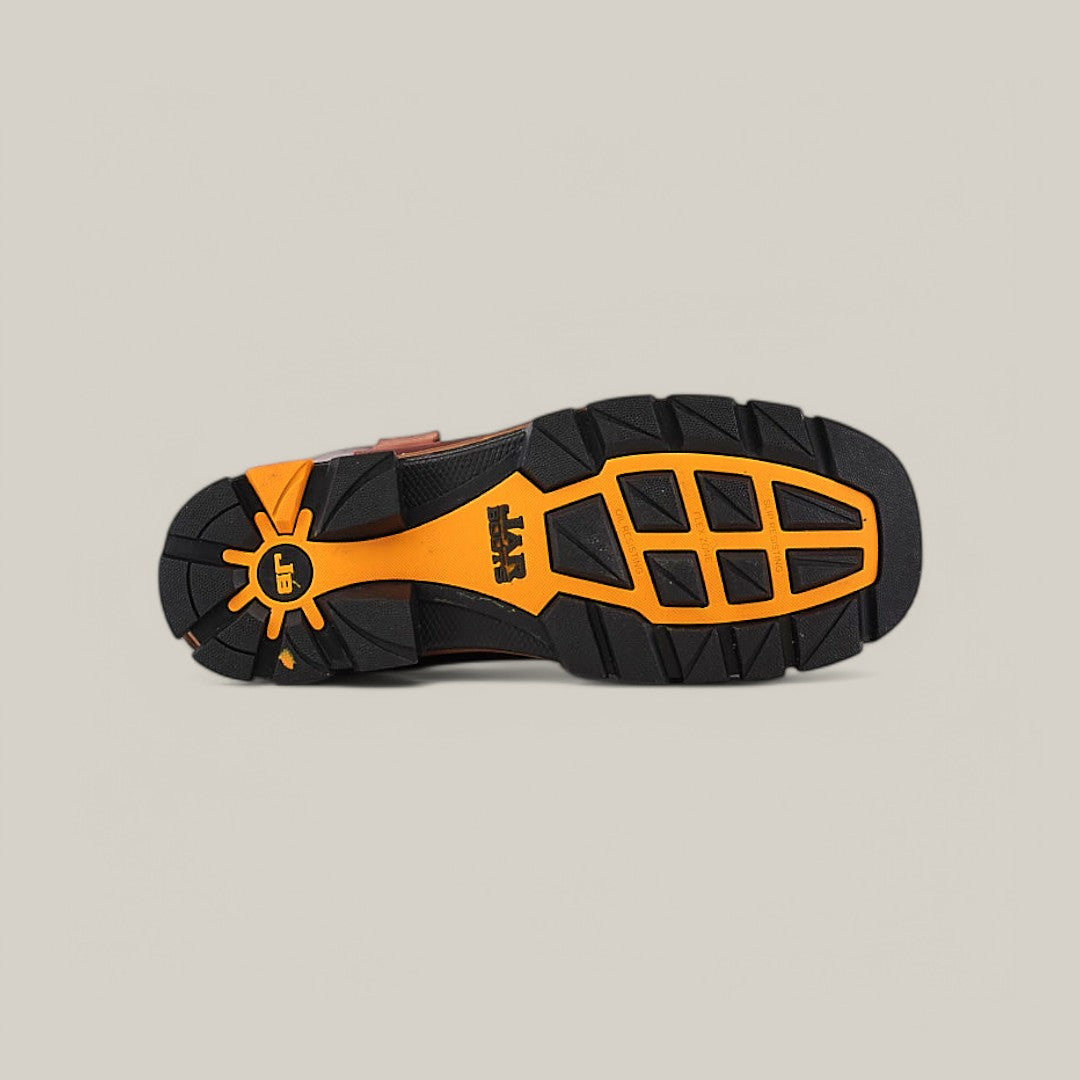 The image displays the sole of an Amazonas Cogñac Composite Toe - E6002 boot, featuring a rugged black design with orange accents. Geometric patterns ensure traction, and a logo is on the heel. Built for durability in tough conditions, its set against a light gray background.