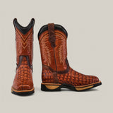 The Alligator Print Cogñac Composite Toe - E6022 boots feature a crocodile skin texture, intricate shaft stitching, black soles, and detailed craftsmanship. Known for durability, these Texas Country Work Boots elegantly blend style with resilience. One boot stands upright; the other shows its side profile.