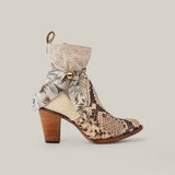 The Plunge Natural Python Zipper & Cut Out - Round Toe - CU570 is a stylish high-heeled boot featuring a textured python design. It boasts a leather strap with metal buckle and floral scarf at the ankle, all complemented by a wooden heel against a plain beige background.