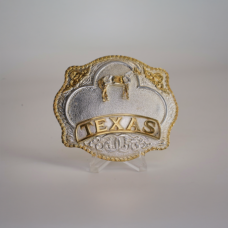 The Texas Horse Buckle boasts a Texas theme with western designs in gold and alpaca silver, featuring a star, horse, and TEXAS embossed in gold. Its elegantly displayed on a clear stand against a plain background.