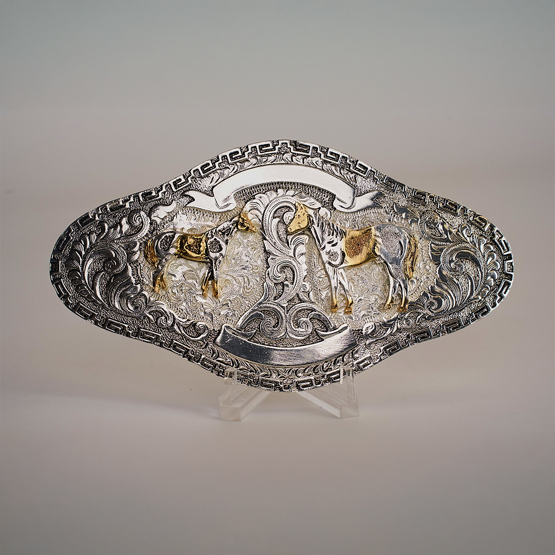 The Long Charro Double Horse Buckle, crafted from alpaca silver, features an ornate, oval design with two gold-toned horses facing each other. The background is adorned with intricate swirls and a border of western geometric patterns. It is elegantly displayed on a clear stand.