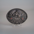 The Ruby Horse Buckle is an alpaca silver oval belt buckle with a raised horse head motif, two red gems, and Texas-style swirls. Its displayed on a clear stand against a neutral background.