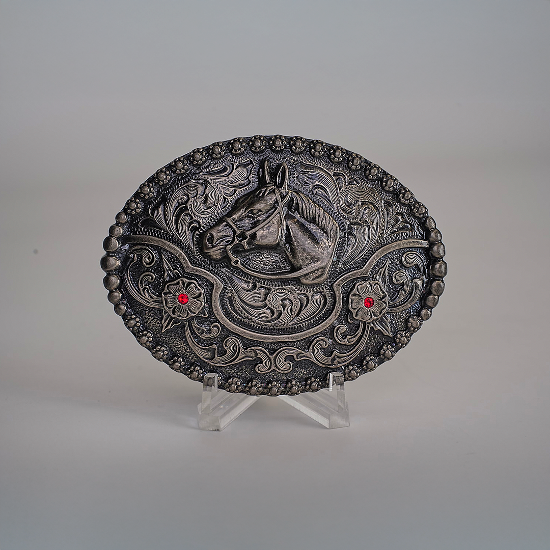 The Ruby Horse Buckle is an alpaca silver oval belt buckle with a raised horse head motif, two red gems, and Texas-style swirls. Its displayed on a clear stand against a neutral background.